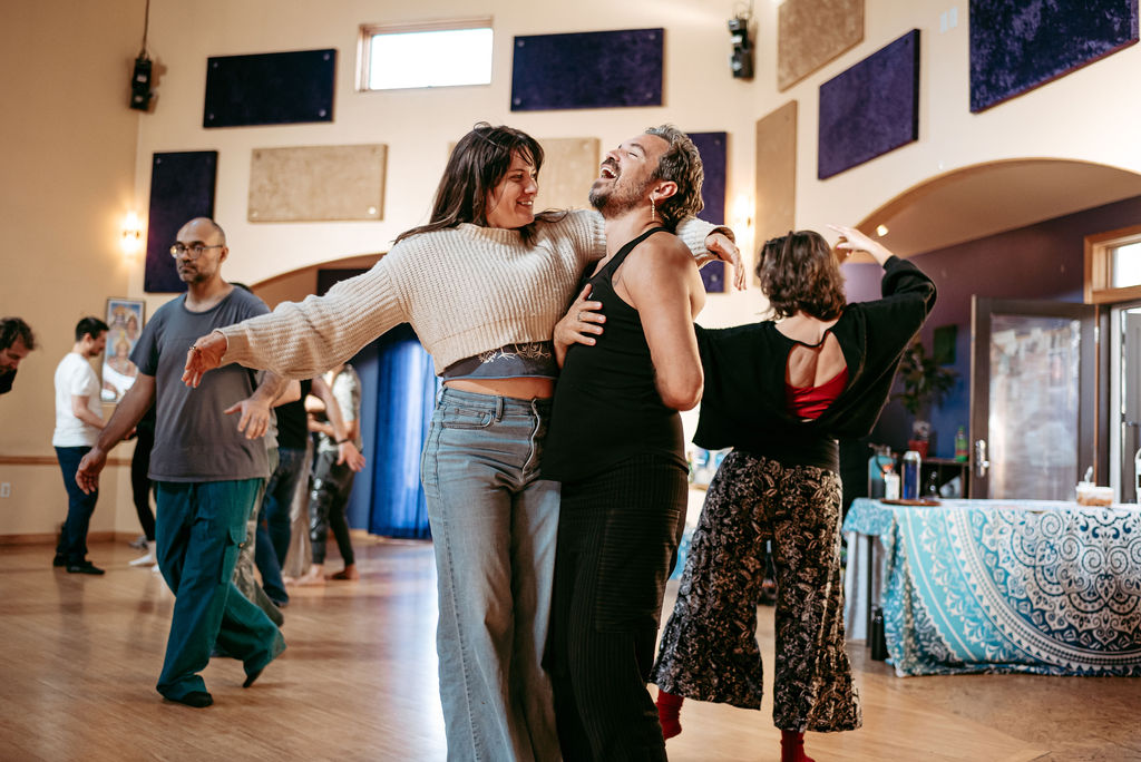 A circle of dancers in flowing earthy clothing moving in a sunlit studio